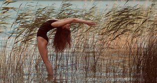  A woman standing in some reeds doing a backbend. The wind is blowing the reeds in the same direction she is bending